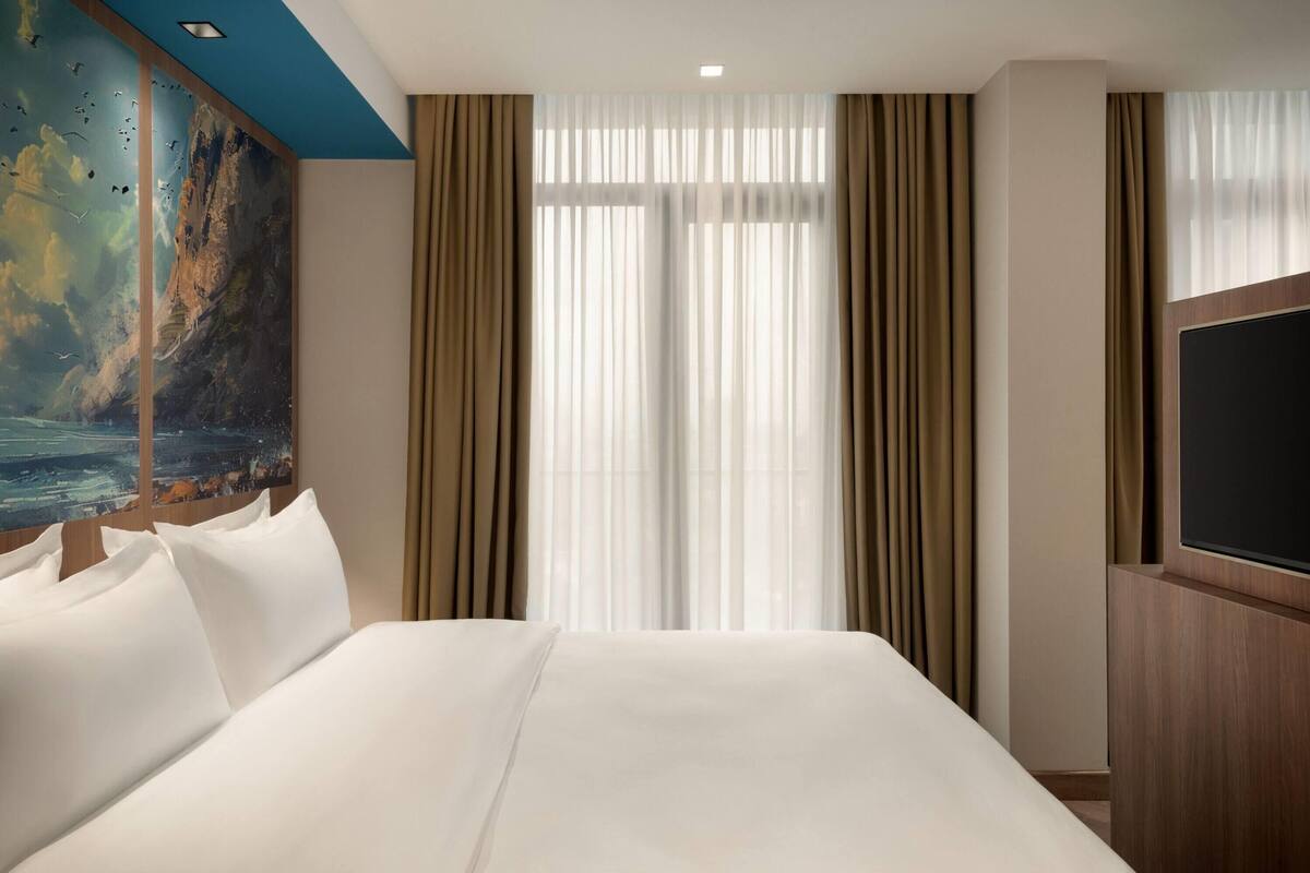 Una camera d'albergo con un letto grande, una televisione a schermo piatto, un dipinto raffigurante una scena marina e tende leggere.