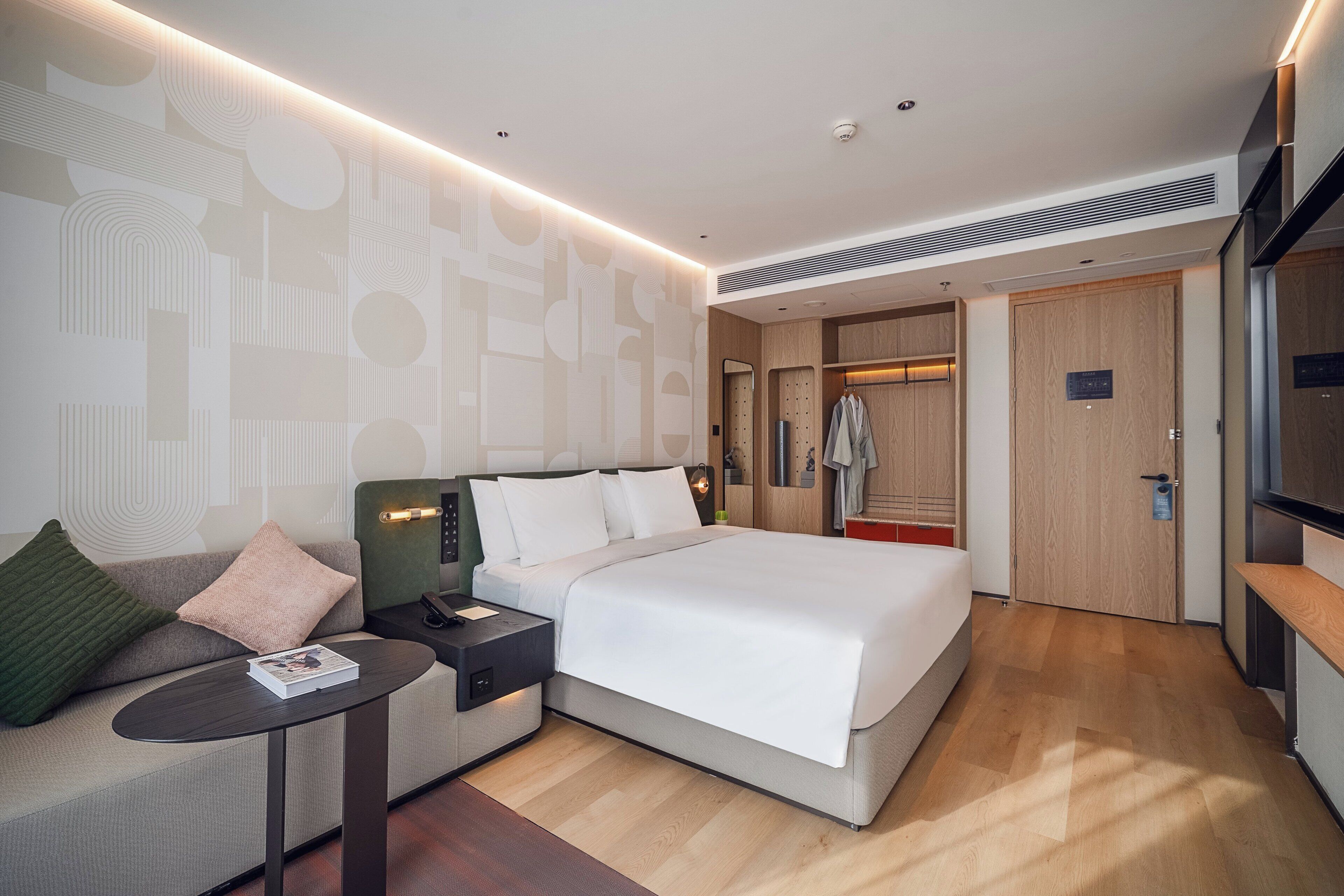 Una habitación de hotel moderna con una cama grande, una mesita de noche, un sofá con cojines, una pared con motivos geométricos y un armario.