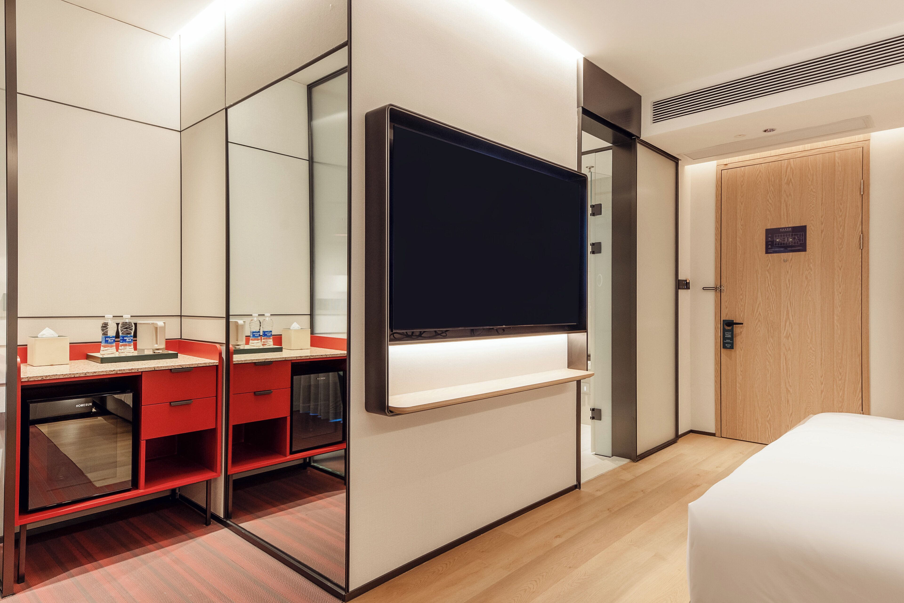 A hotel room with a large mirror, a flat-screen TV, a wooden door, and a bed.
