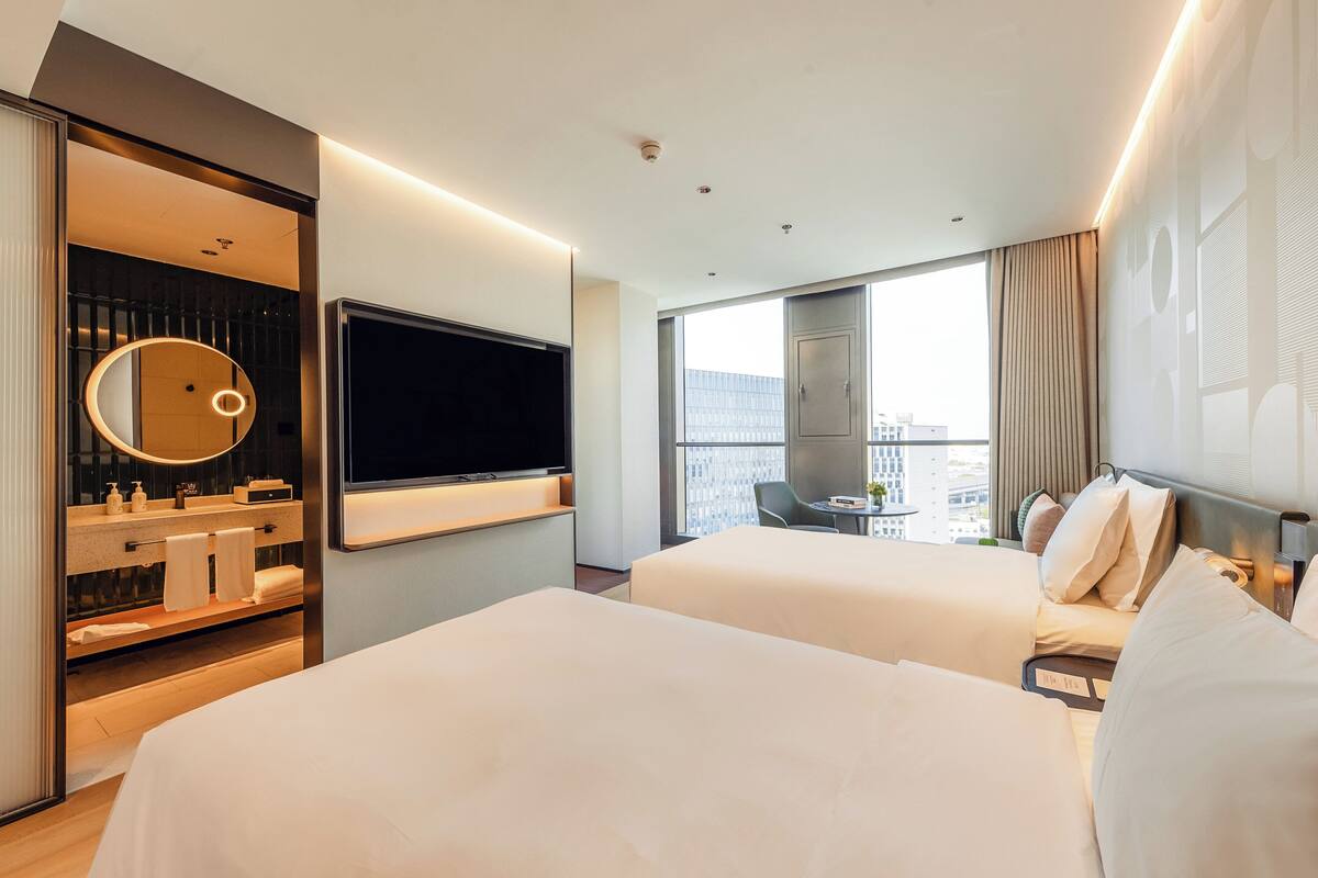 A hotel room with two beds, a TV, a bathroom with a mirror, and a city view.