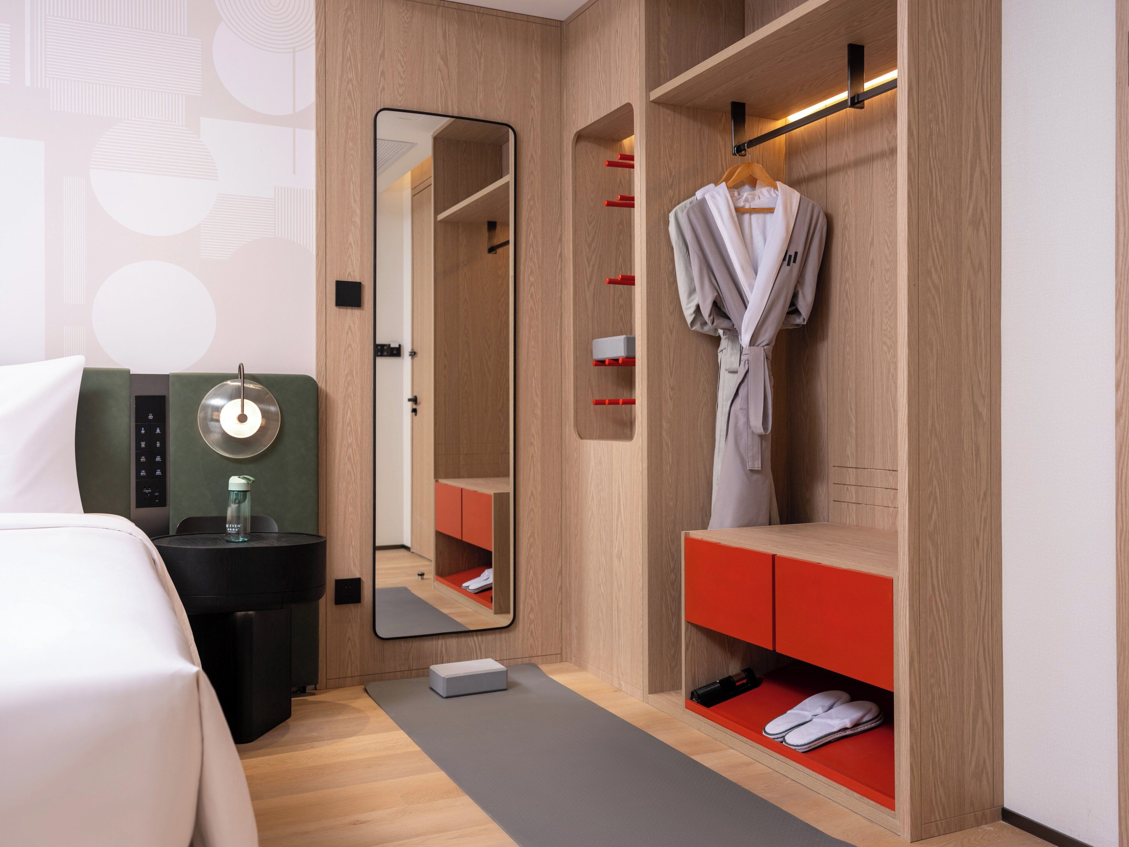A hotel room with a bed, a mirror, a robe hanging, a red storage unit, and a small exercise mat.