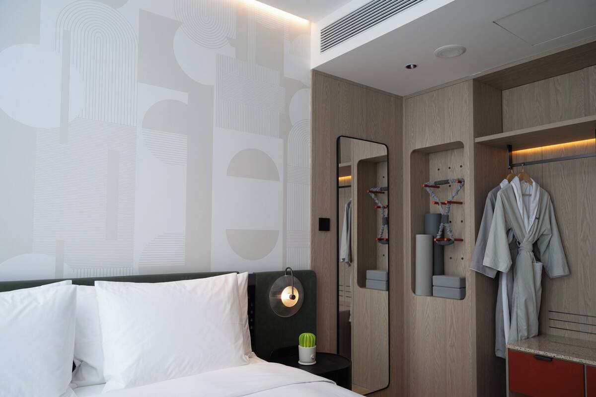 A modern hotel room with a large bed, a bedside table with a lamp, a mirror, a wooden wardrobe, and a red cabinet with a shoe rack.