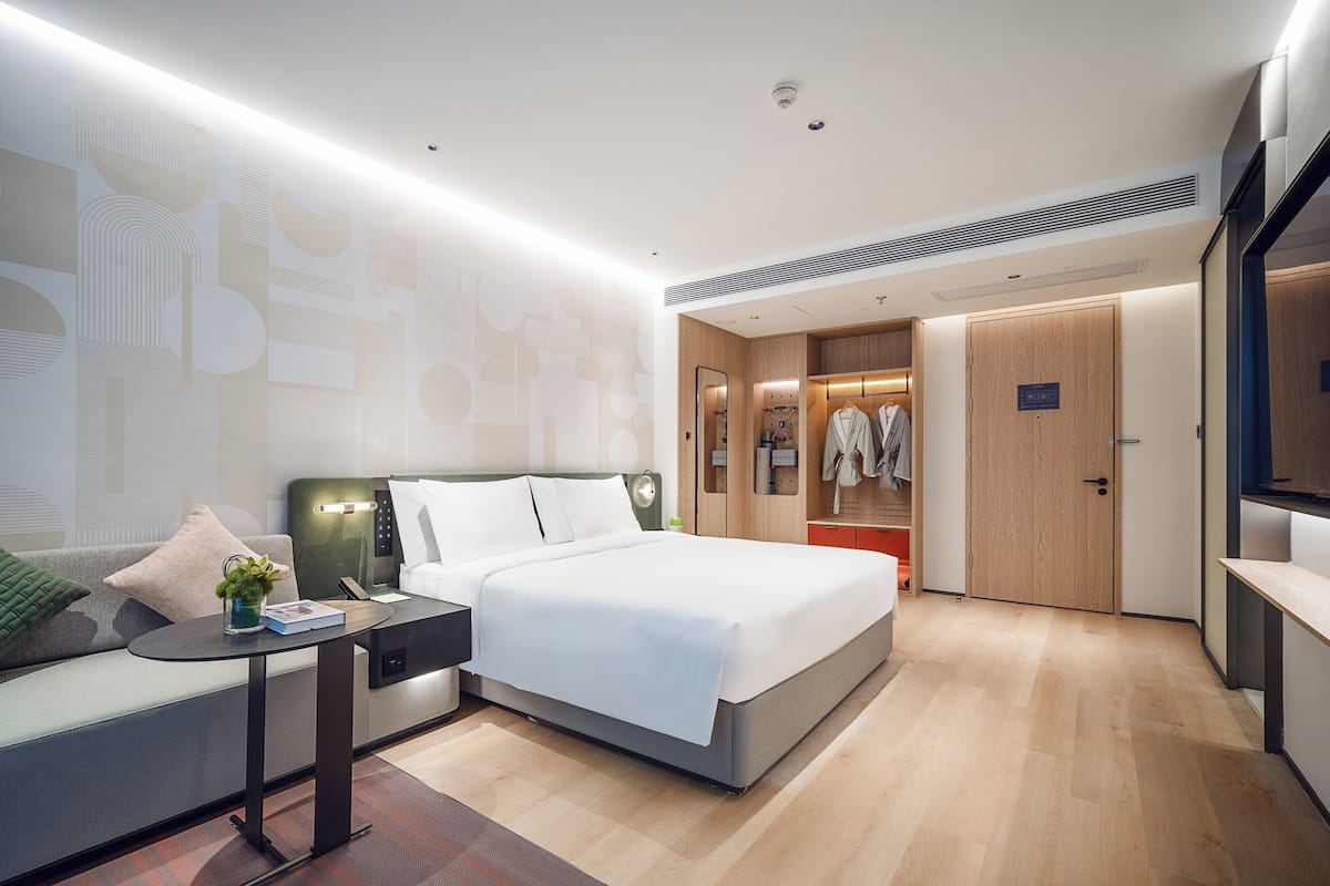 A modern hotel room with a neatly made bed, a seating area with a small table, and a wardrobe with a mirror.