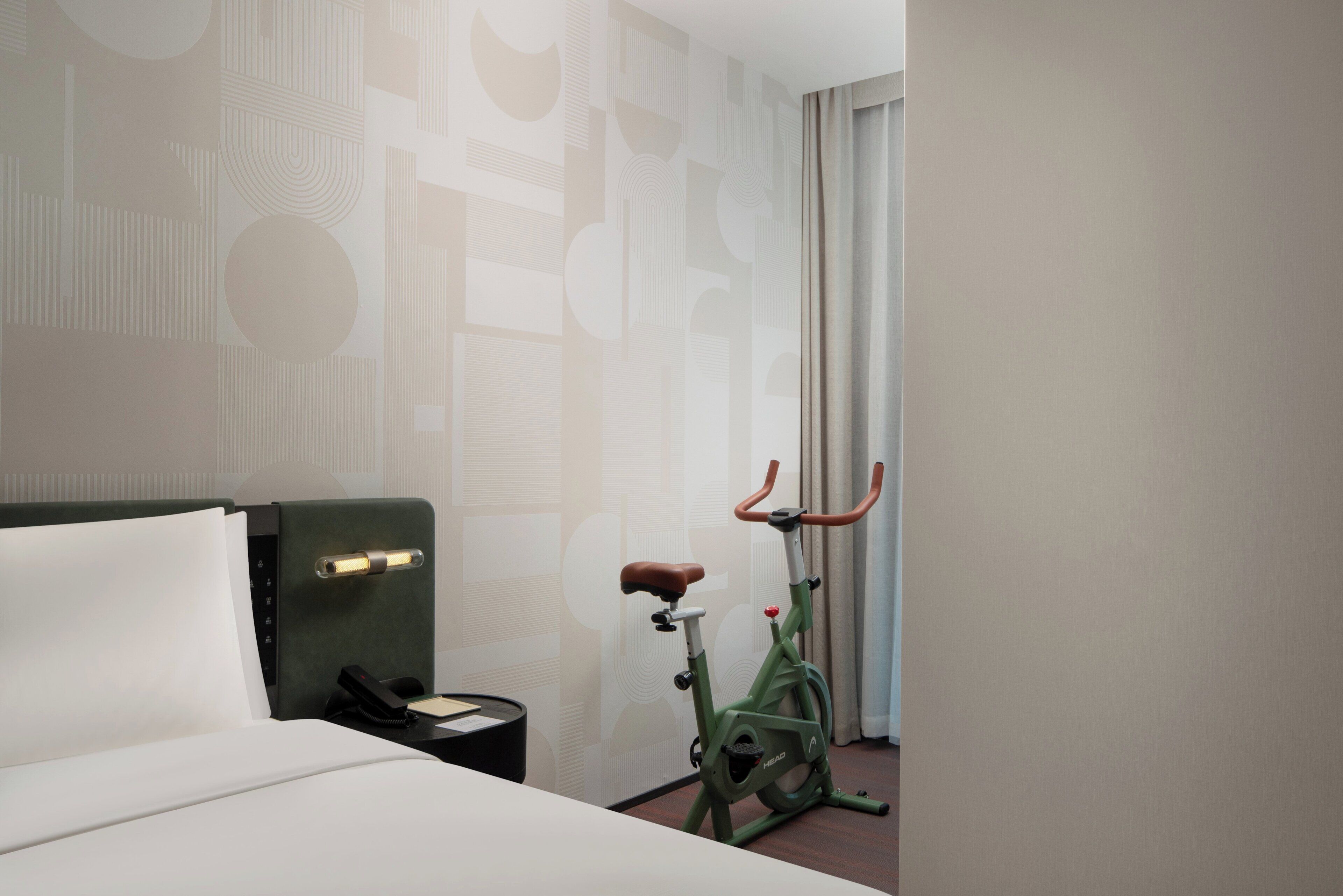Quarto de hotel com cama, uma bicicleta ergométrica verde, mesa de cabeceira com telefone e parede estampada.