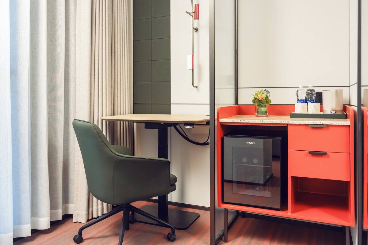 A hotel room with a desk, chair, minibar, and a small plant on the desk.
