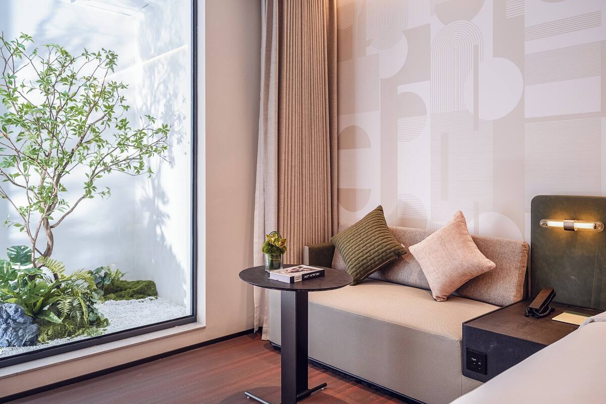 A modern hotel room with a large window, a small round table with a book and a plant, a sofa with cushions, and a bedside lamp.
