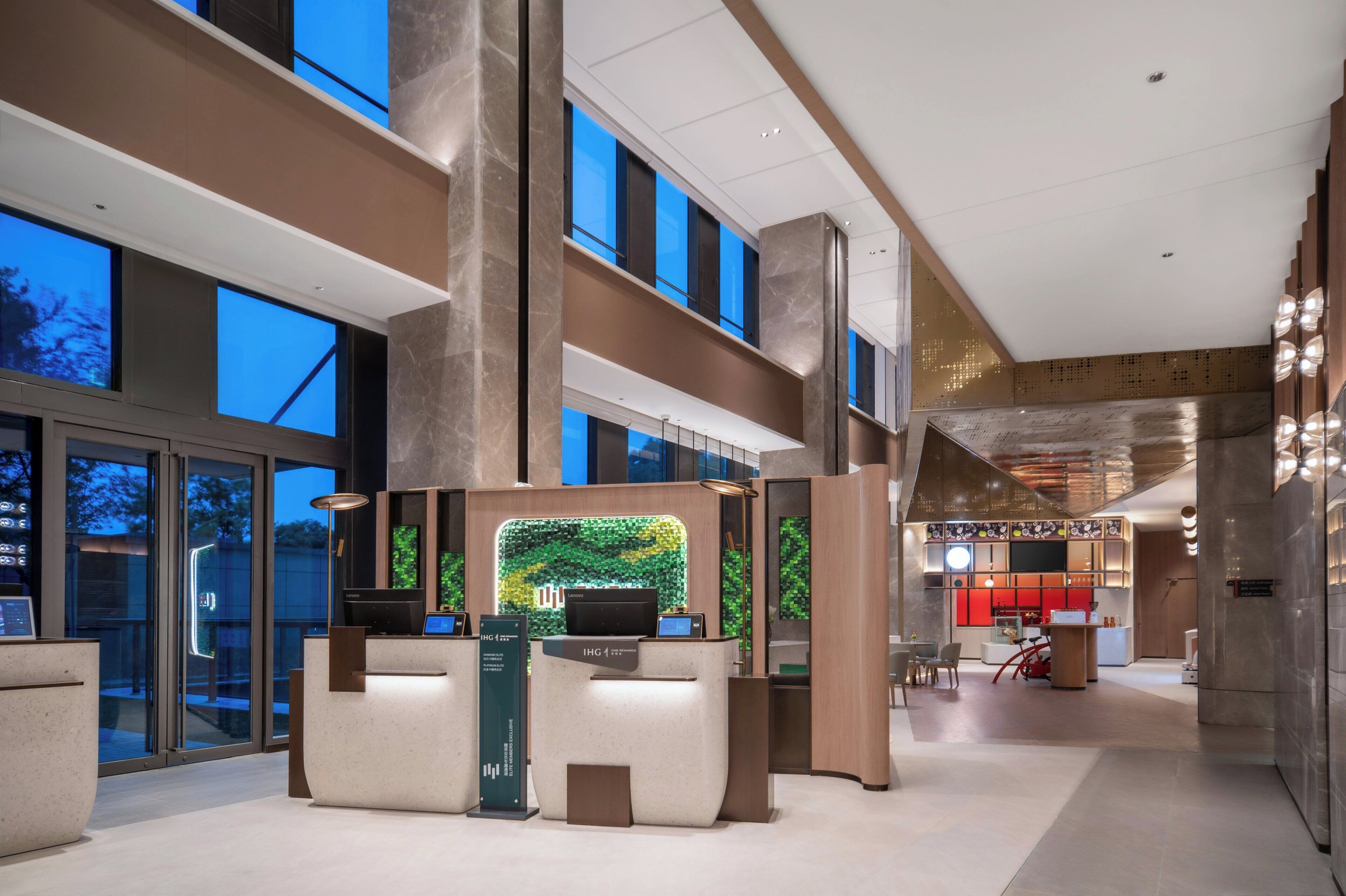 A modern hotel lobby with a reception desk, a green wall feature, and a dining area in the background.