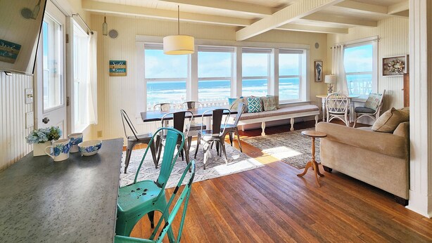 A coastal living room with a wooden ceiling, large windows overlooking the ocean, a dining area with chairs and a table, and a cozy seating area with a sofa and armchairs.