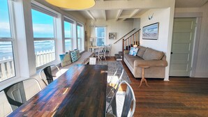 A spacious living room with a large wooden dining table, a sofa, and a view of the ocean through large windows.