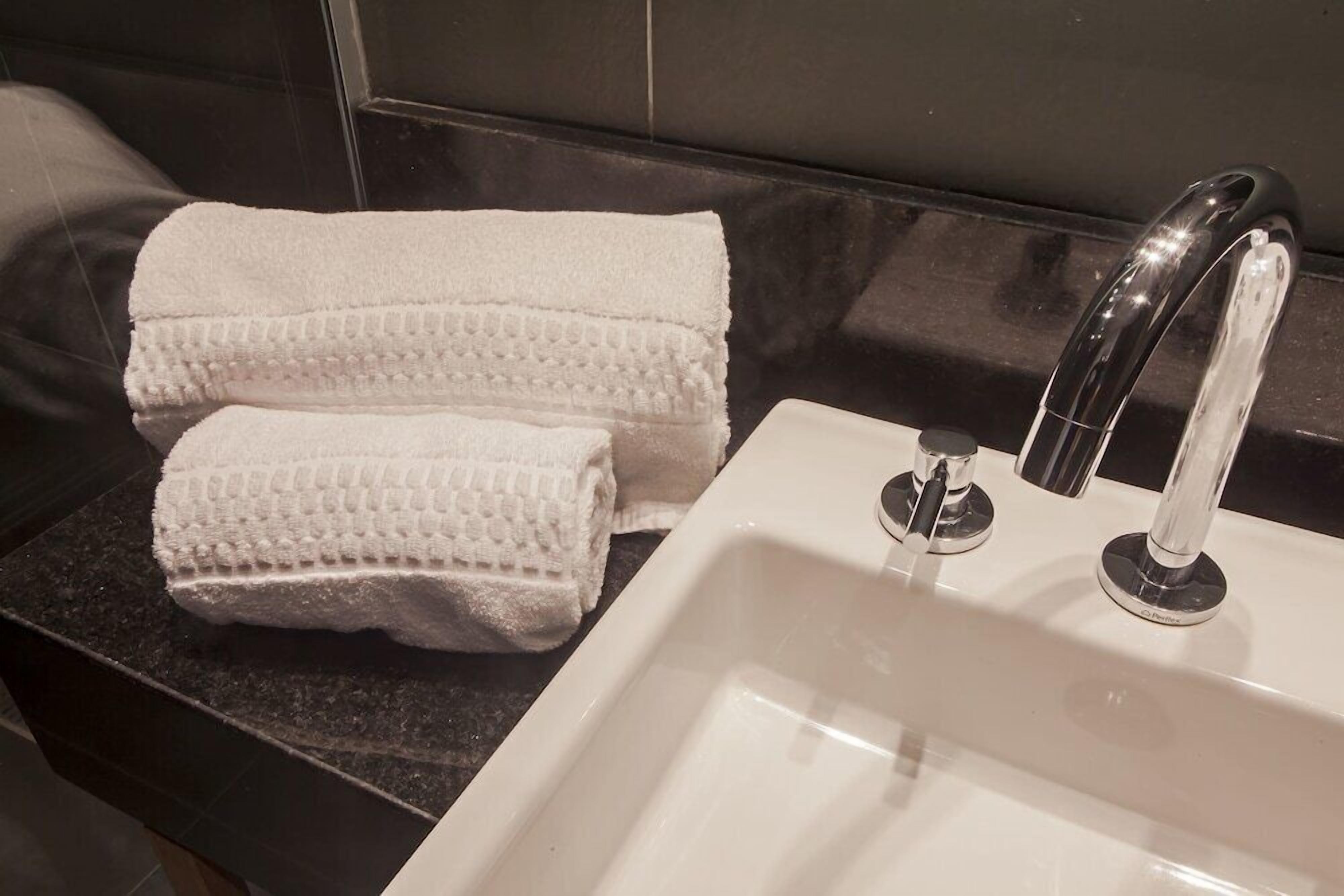 Un baño con un lavamanos blanco, un grifo moderno y dos toallas enrolladas sobre una encimera oscura.