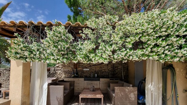 An outdoor patio with wicker furniture, a stone wall, and a pergola covered with flowering vines. - Jardin d'Uzès walking distance village (Saint-Quentin-la-Poterie)