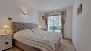 A bedroom with a large bed, a nightstand with a lamp, a window with curtains, and a wall-mounted picture frame. - Jardin d'Uzès walking distance village (Saint-Quentin-la-Poterie)