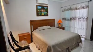 A bedroom with a bed, a nightstand, a chair, a lamp, a painting, and a window with curtains. - Casa Real Hotel (Degollado)