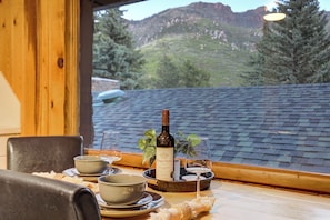 A dining table with plates, bowls, a bottle of wine, and glasses, set against a window overlooking a mountainous landscape.