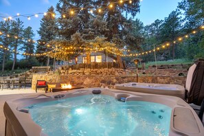 A hot tub area with a stone wall and wooden deck, surrounded by trees and string lights. - The Stone Lodge | Sleeps 14 | Hot Tubs | Game Room (Cascade)