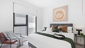A modern bedroom with a large bed, a chair, a bedside table, and a window with blinds. - Urban Living at Its Best (Carnegie)