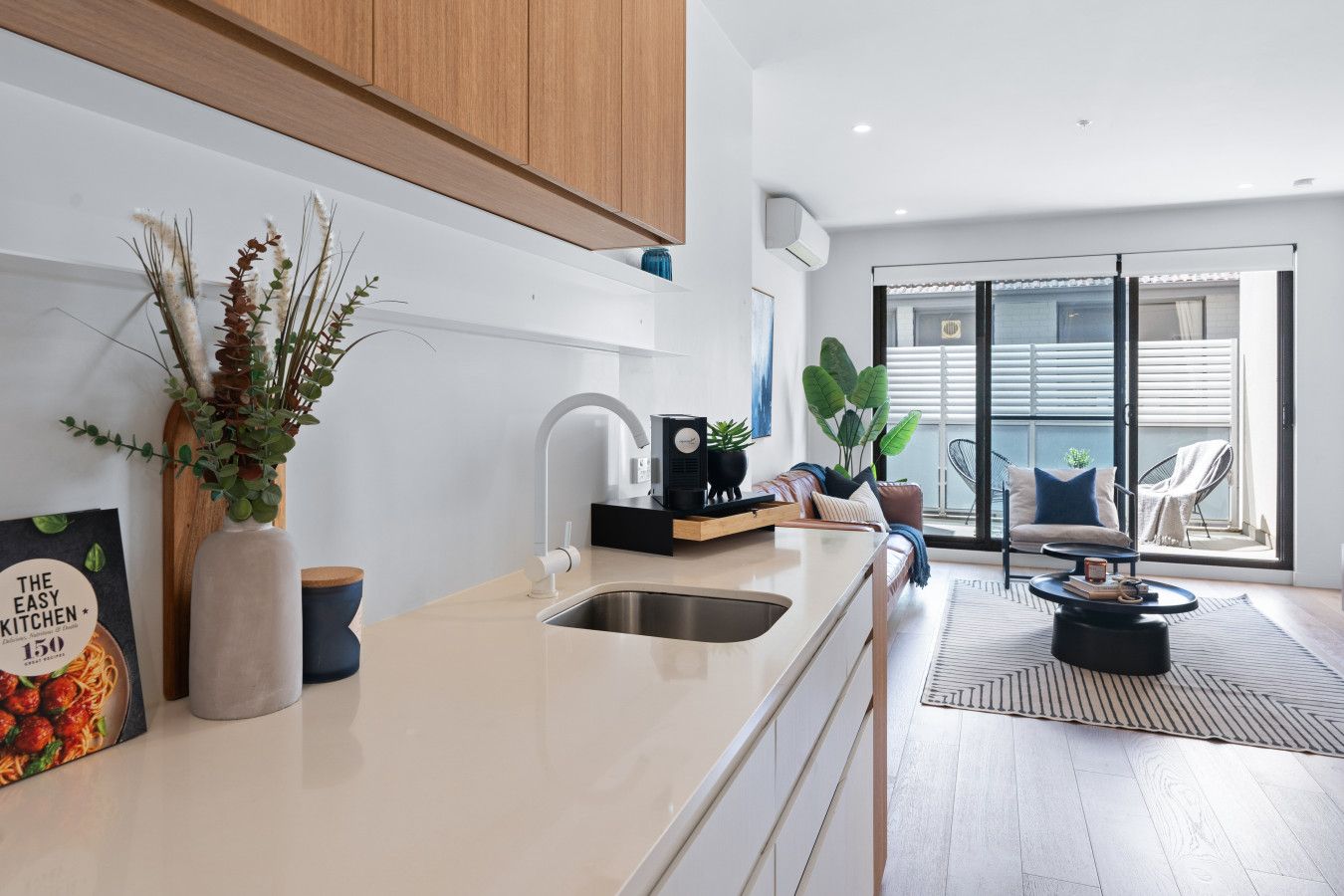 A modern kitchen with a white countertop, stainless steel sink, and wooden cabinets. There is a dining area with a black chair and a round table.