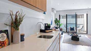 A modern kitchen with a white countertop, stainless steel sink, and wooden cabinets. There is a dining area with a black chair and a round table.