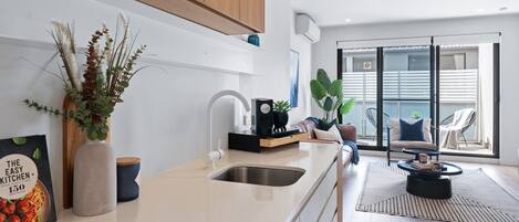 A modern kitchen with a white countertop, stainless steel sink, and wooden cabinets. There is a dining area with a black chair and a round table.