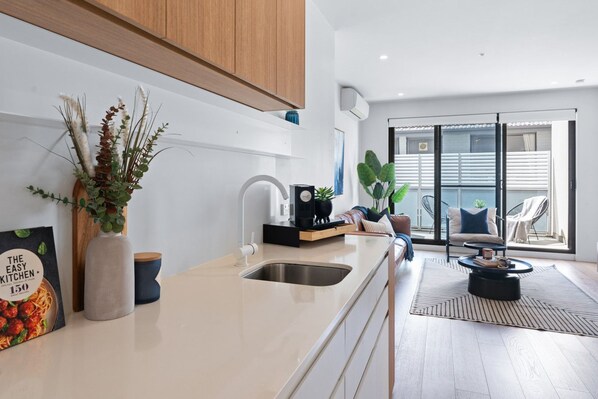 A modern kitchen with a white countertop, stainless steel sink, and wooden cabinets. There is a dining area with a black chair and a round table.