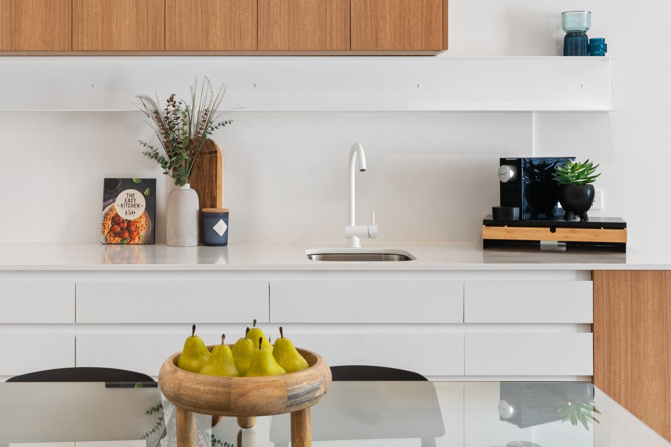 Une cuisine moderne avec des meubles blancs, un évier en acier inoxydable et un panier à fruits en bois posé sur une table en verre.