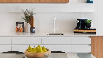 Une cuisine moderne avec des meubles blancs, un évier en acier inoxydable et un panier à fruits en bois posé sur une table en verre.