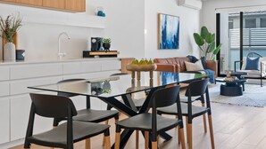 Et moderne køkken med hvide skabe, et glasbord til spisning og sorte stole. Der er en sofa og et sofabord i opholdsstuen, samt en stor plante i hjørnet.