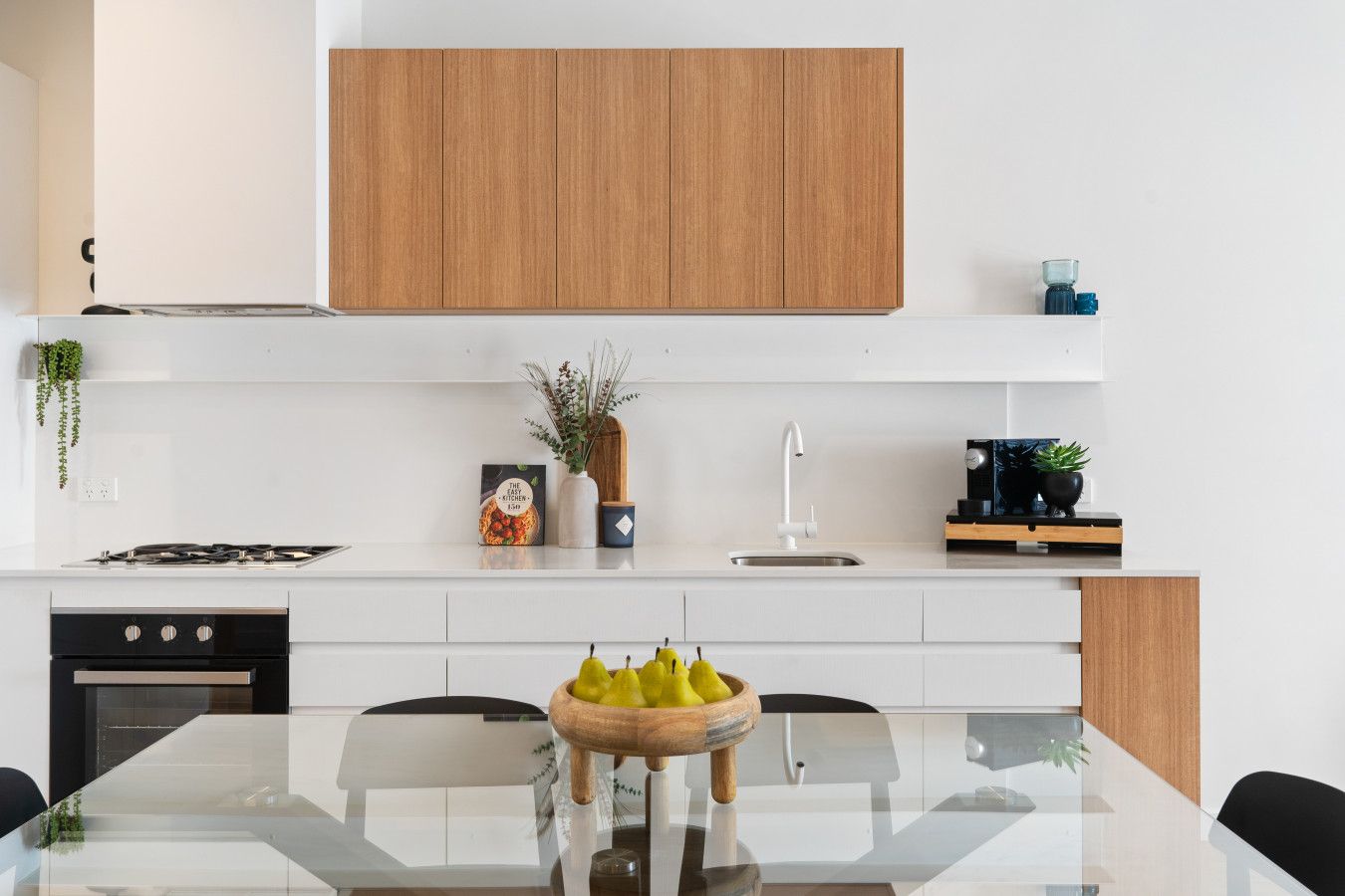 Une cuisine moderne avec des meubles blancs, une table à manger en verre et un saladier en bois rempli de fruits.