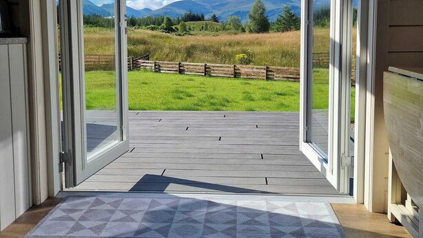 An open doorway leading to a deck with a view of a grassy field and mountains. - Toradh Cabin (Spean Bridge)
