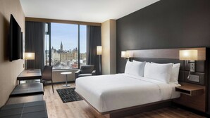A modern hotel room with a large bed, a desk, a chair, and a TV. The room has a city view through large windows. - AC Hotel BY Marriott Ottawa Dwtn (OTTAWA)
