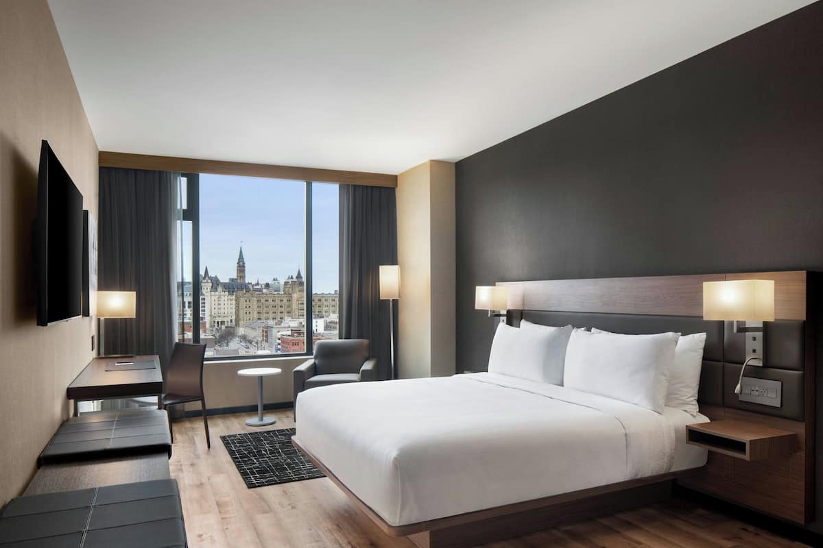 Habitación de hotel moderna con una cama grande, un escritorio, una silla y un televisor. La habitación tiene vistas a la ciudad a través de amplias ventanas.