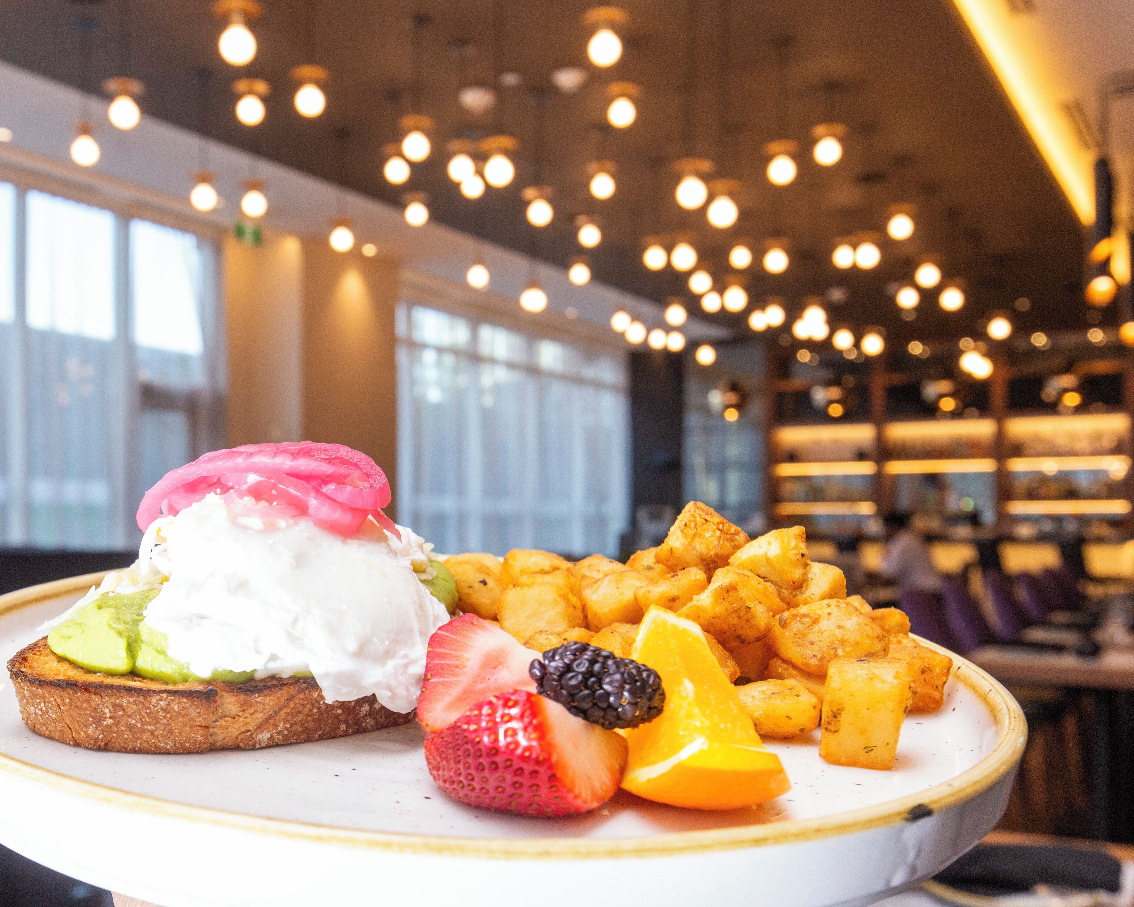 Een bord met toast belegd met avocado, slagroom en bessen, met gebakken aardappelpartjes erbij.