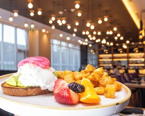 Une assiette avec des tranches de pain grillé garnies d’avocat, de crème fouettée et de baies, accompagnée de morceaux de pommes de terre frits.