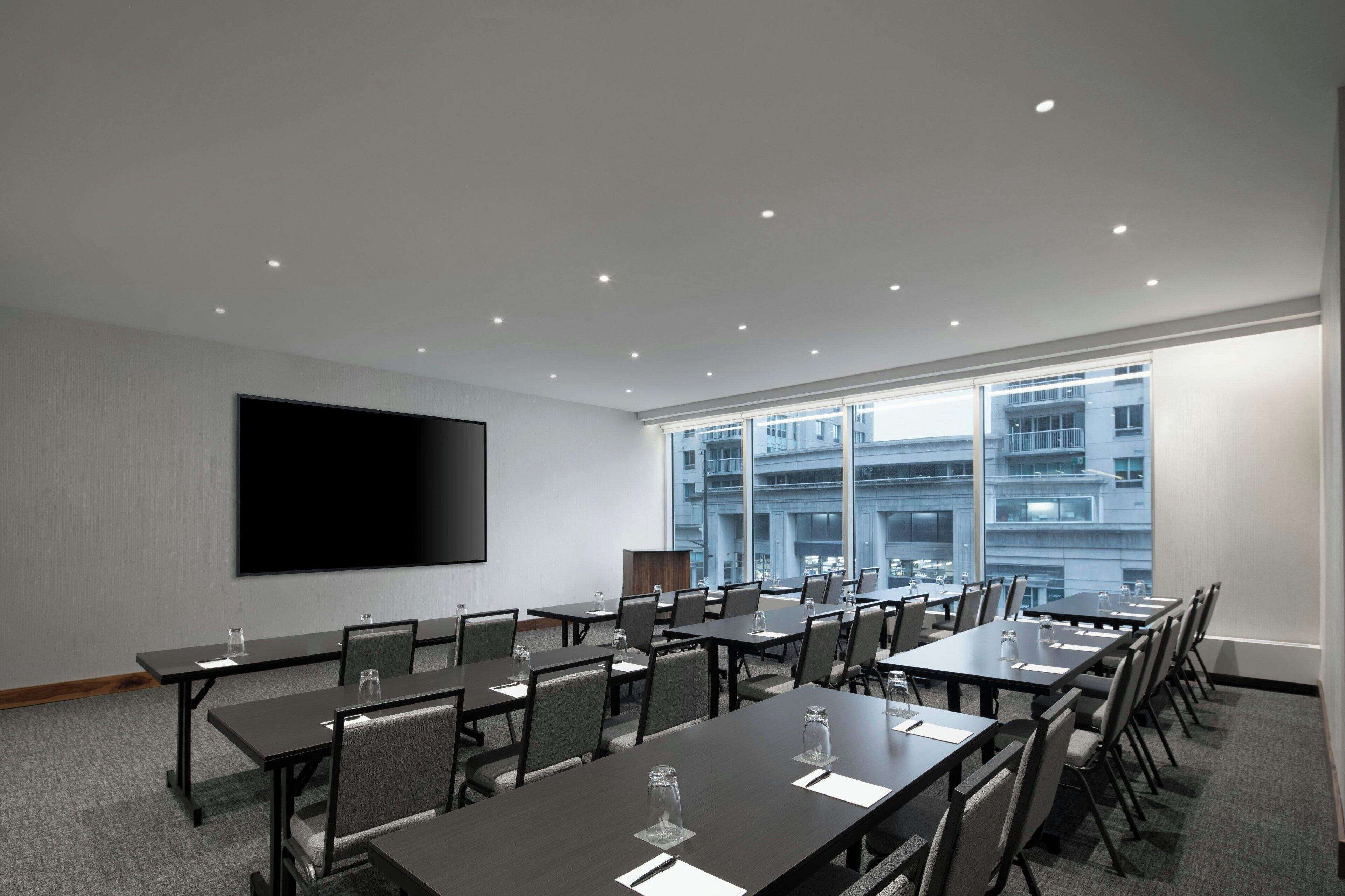 A conference room with multiple tables and chairs, a large flat-screen TV on the wall, and large windows with a city view.