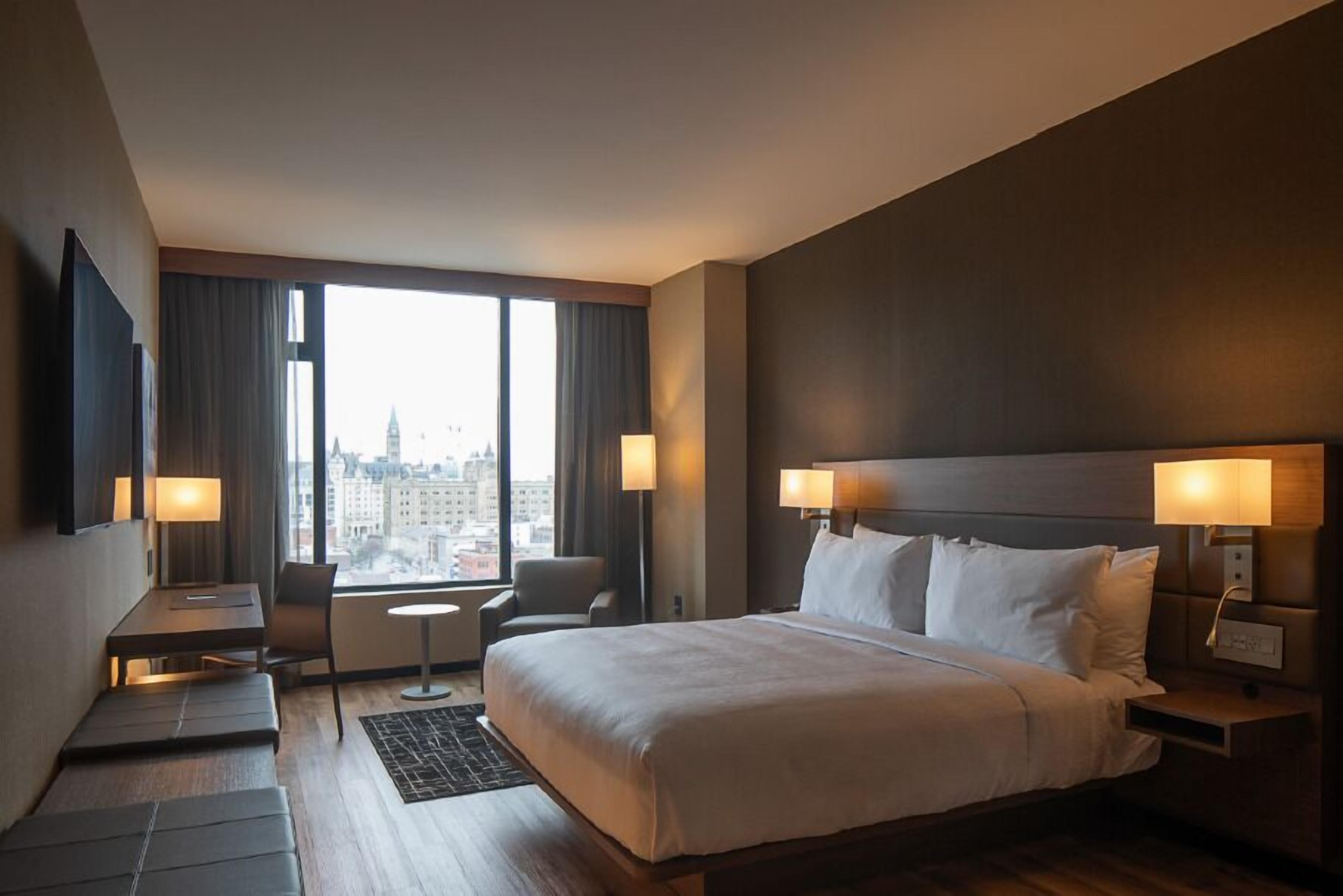 A hotel room with a large bed, two bedside tables with lamps, a desk with a chair, and a view of the city through large windows.
