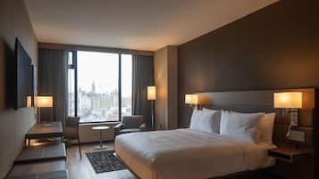 A hotel room with a large bed, two bedside tables with lamps, a desk with a chair, and a view of the city through large windows.