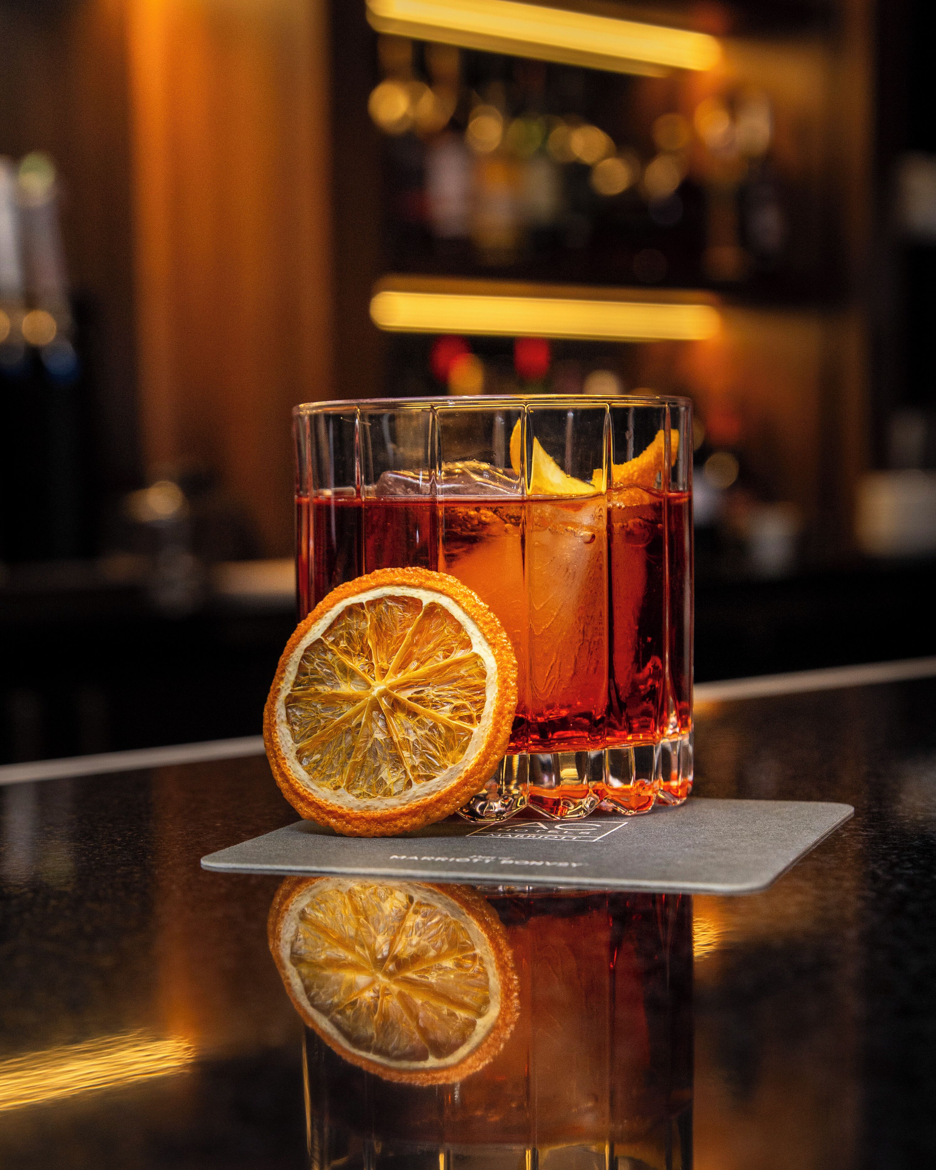 A glass of amber-colored beverage with a slice of dried orange garnish on a reflective bar counter.