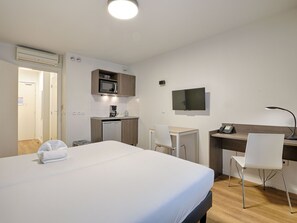A hotel room with a bed, a desk, a chair, a TV, and a kitchenette.