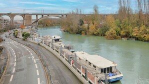 A river with a bridge in the background, a boat docked along the riverbank, and a road with directional arrows. - Cosy studio proche RER, idéal pour visiter Paris (Nogent-sur-Marne)