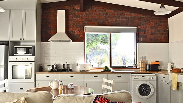 A modern kitchen with white cabinets, a brick wall, a window, and a built-in washing machine. - UzuriBarossa, Tranquil guesthouse retreat in Lyndoch, Barossa Valley (Altona)