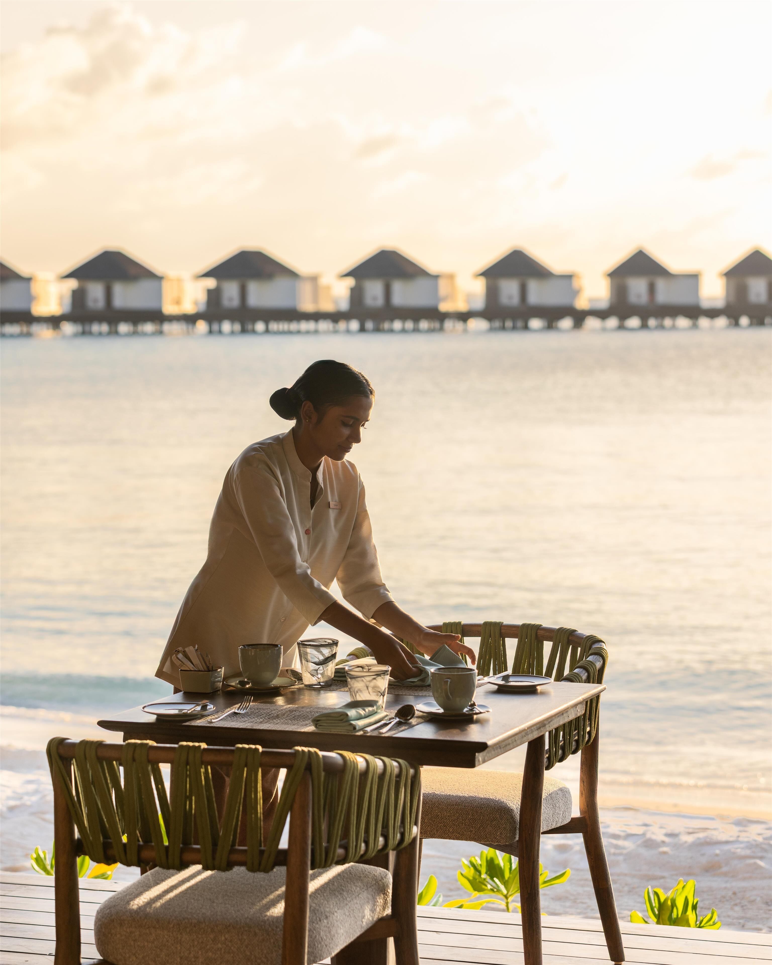 A person setting a table outdoors with a view of overwater bungalows in the background.