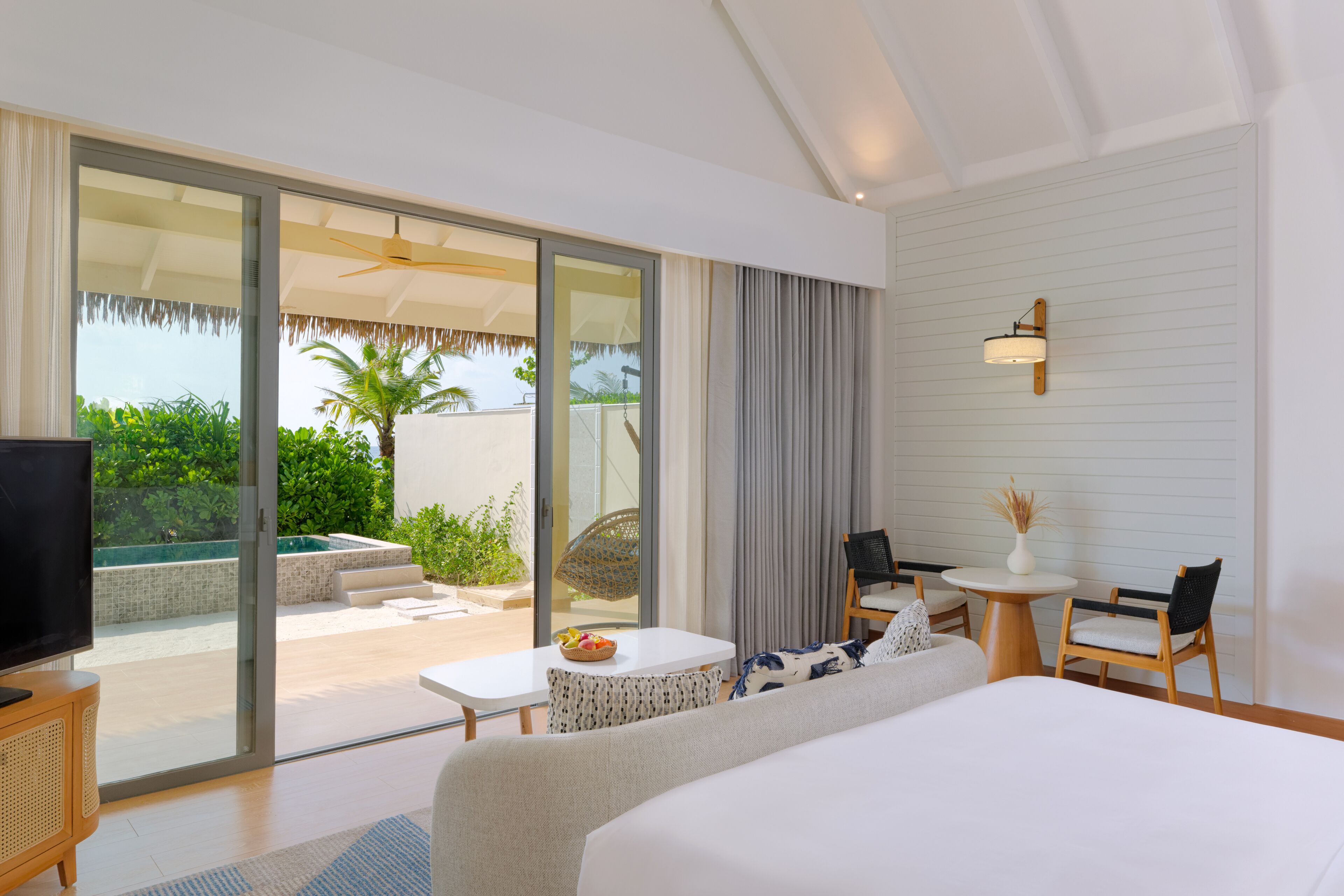 Quarto de hotel moderno com uma cama grande, televisão, área de refeições com mesa e cadeiras, e porta de vidro deslizante que dá para uma área de piscina externa.