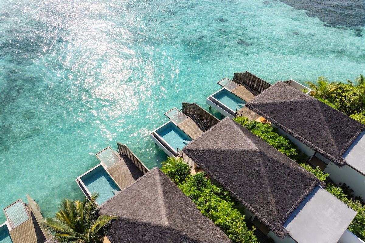 Vue aérienne d’un complexe hôtelier avec des bungalows sur pilotis, une eau turquoise cristalline et une végétation luxuriante.
