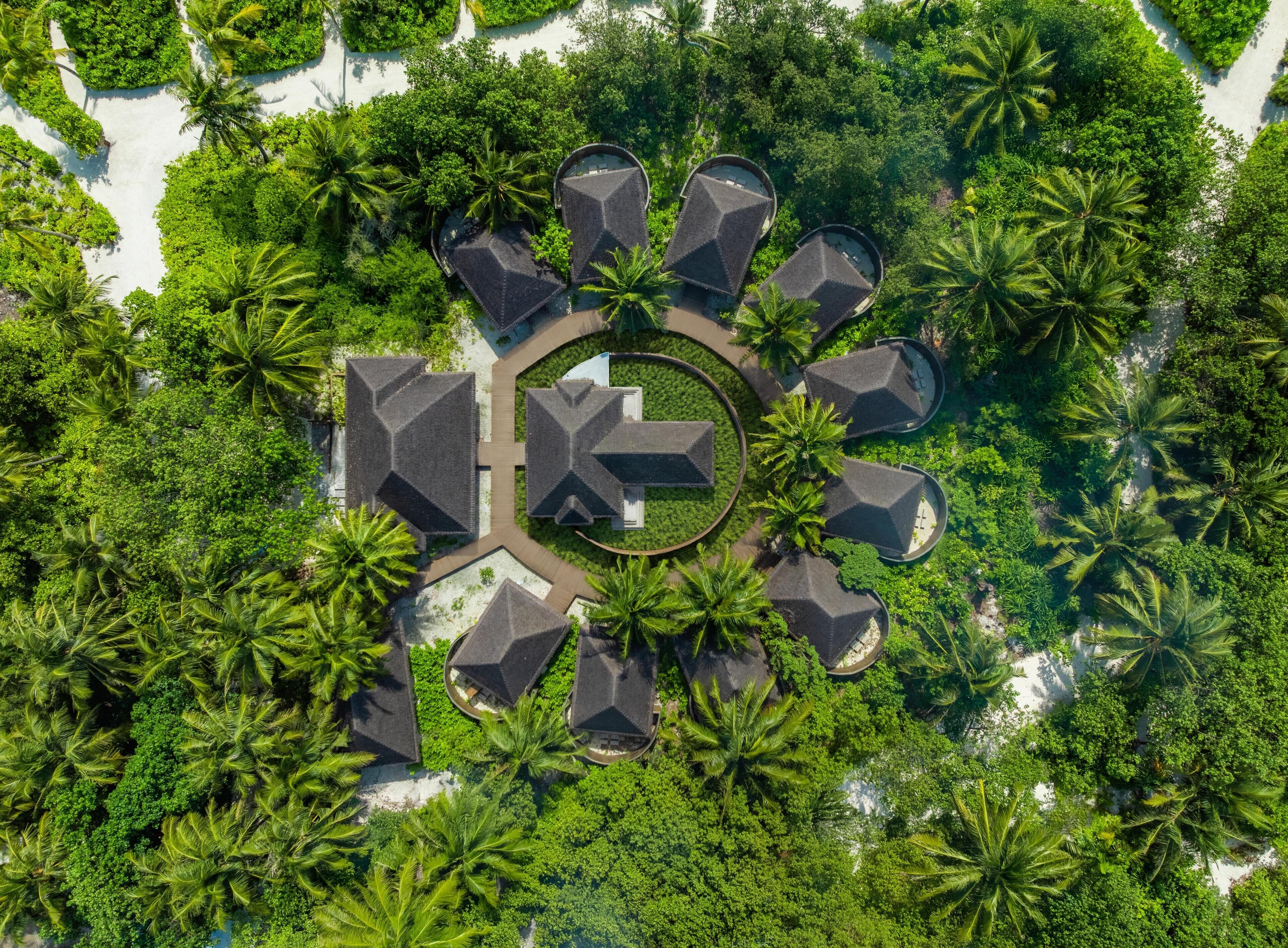 Une vue aérienne d’un complexe hôtelier composé de plusieurs bâtiments de style bungalow, disposés en cercle autour d’une cour centrale, le tout entouré d’une végétation luxuriante et de palmiers.