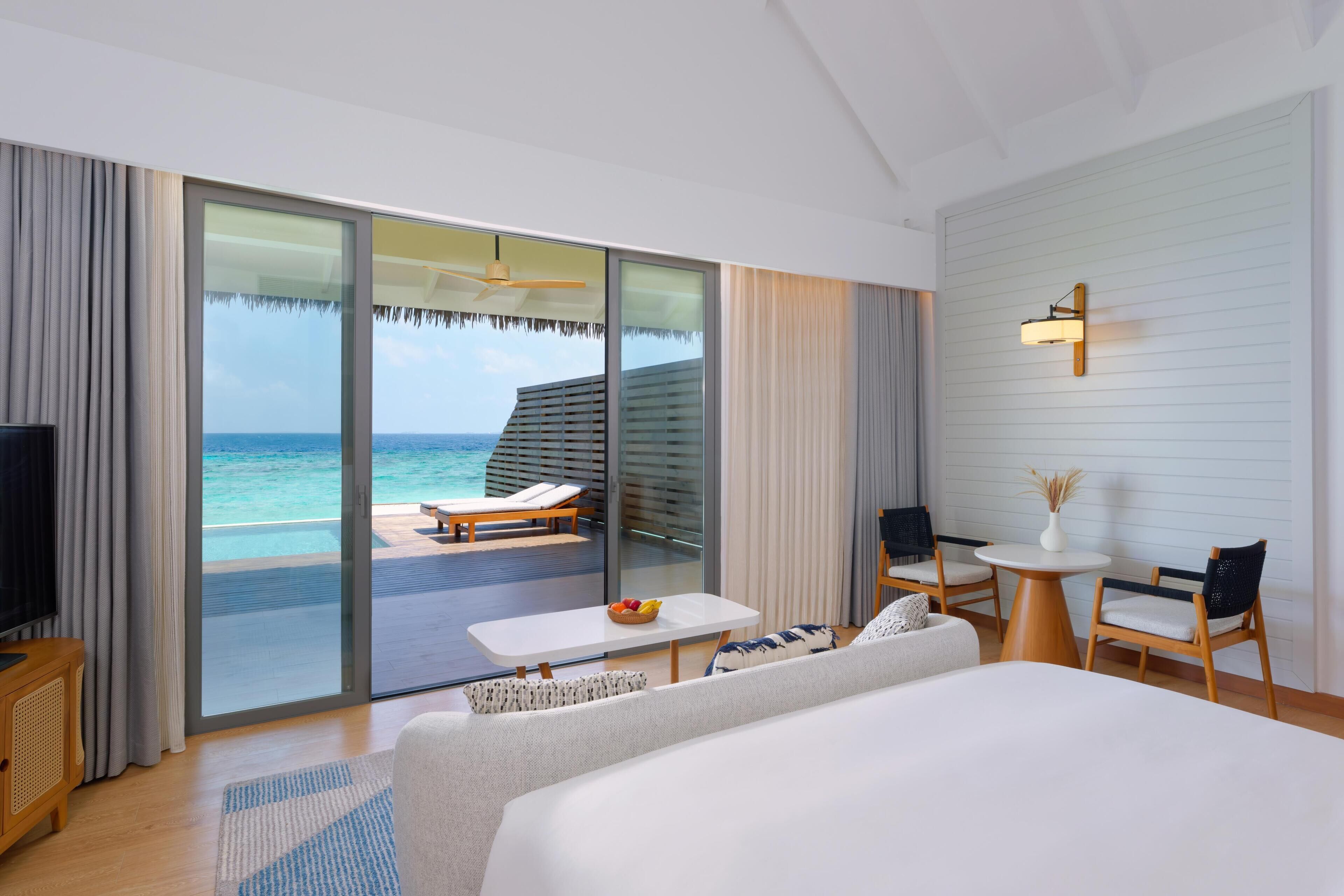 A modern hotel room with a large bed, a TV, a dining area with a table and chairs, and a balcony with a view of the ocean.