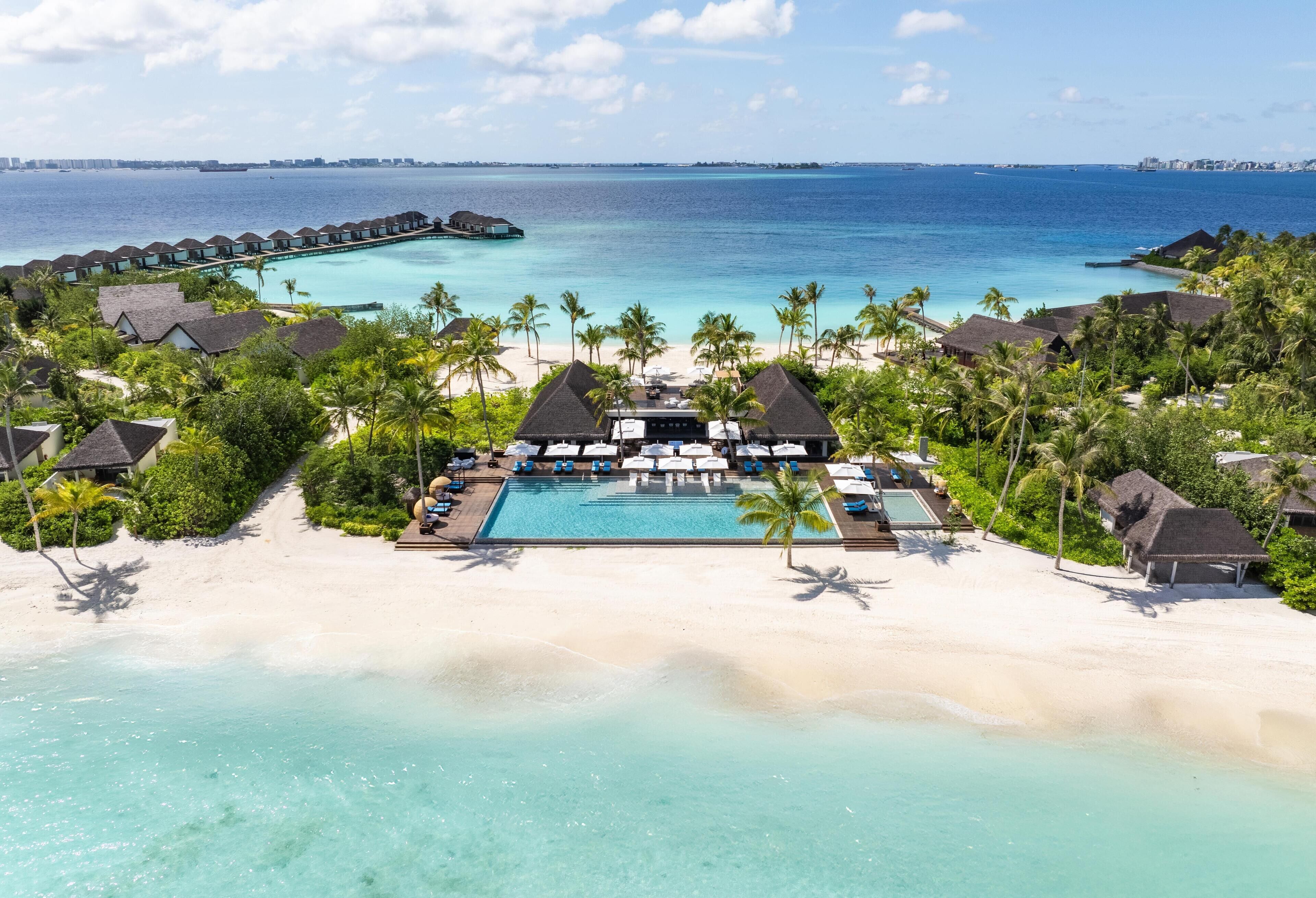An aerial view of a resort with overwater bungalows, a swimming pool, and a beach.