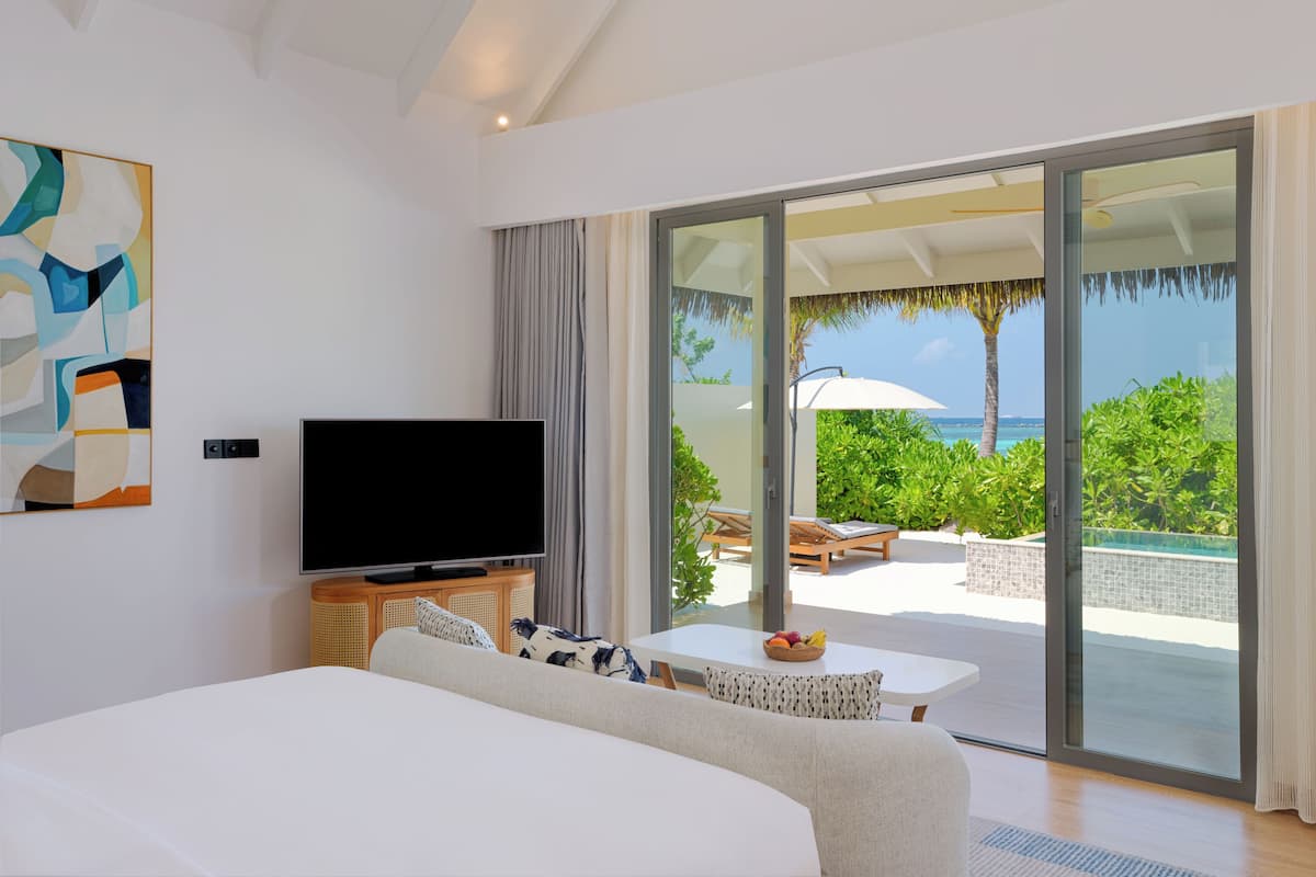 Une chambre d’hôtel moderne dotée d’un grand lit, d’un téléviseur à écran plat, d’une table à manger avec un panier de fruits, et d’une porte-fenêtre coulissante donnant sur un espace piscine aménagé avec du mobilier d’extérieur.