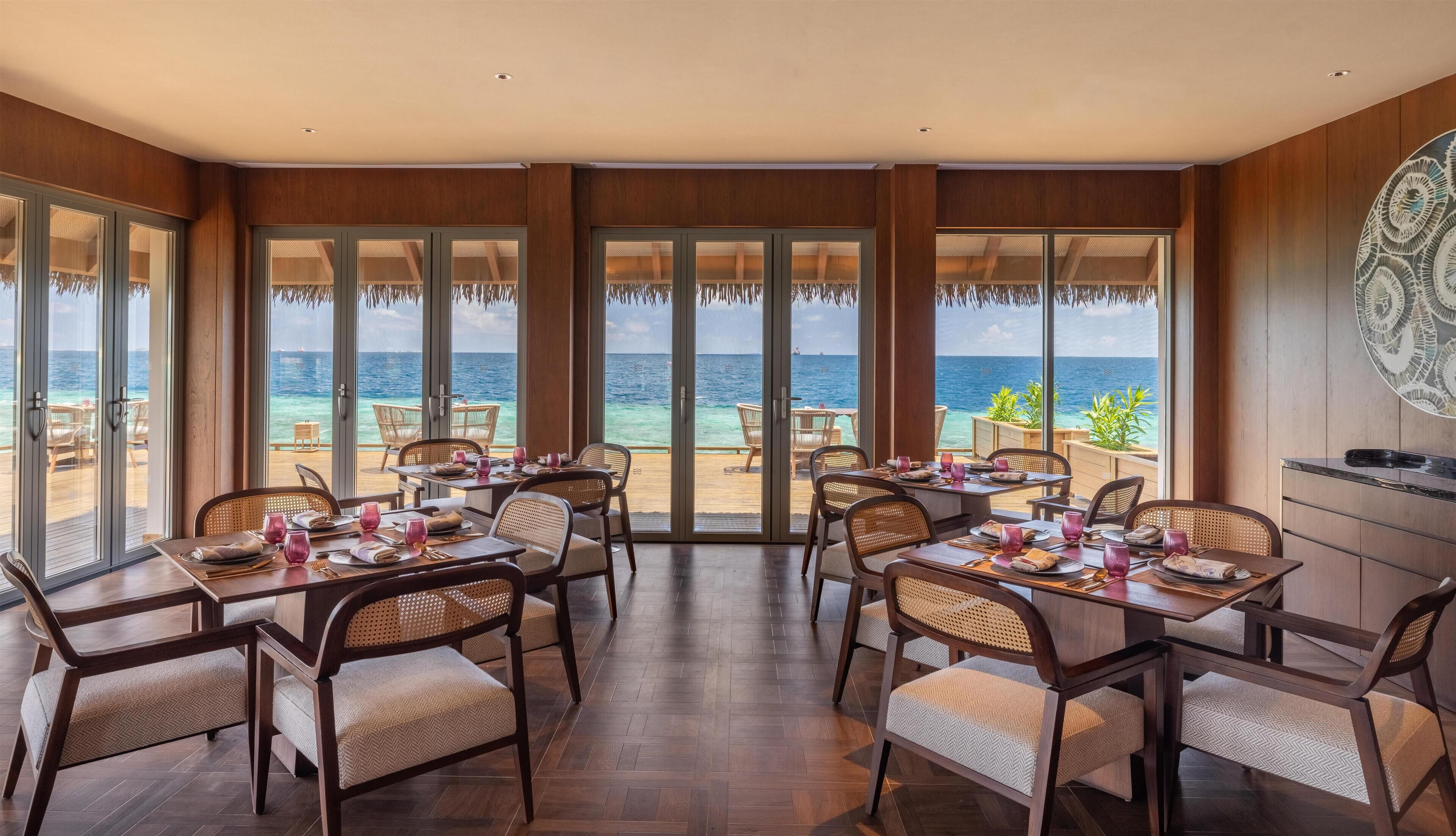 A dining area with tables and chairs, large windows with ocean views, and a wooden interior.