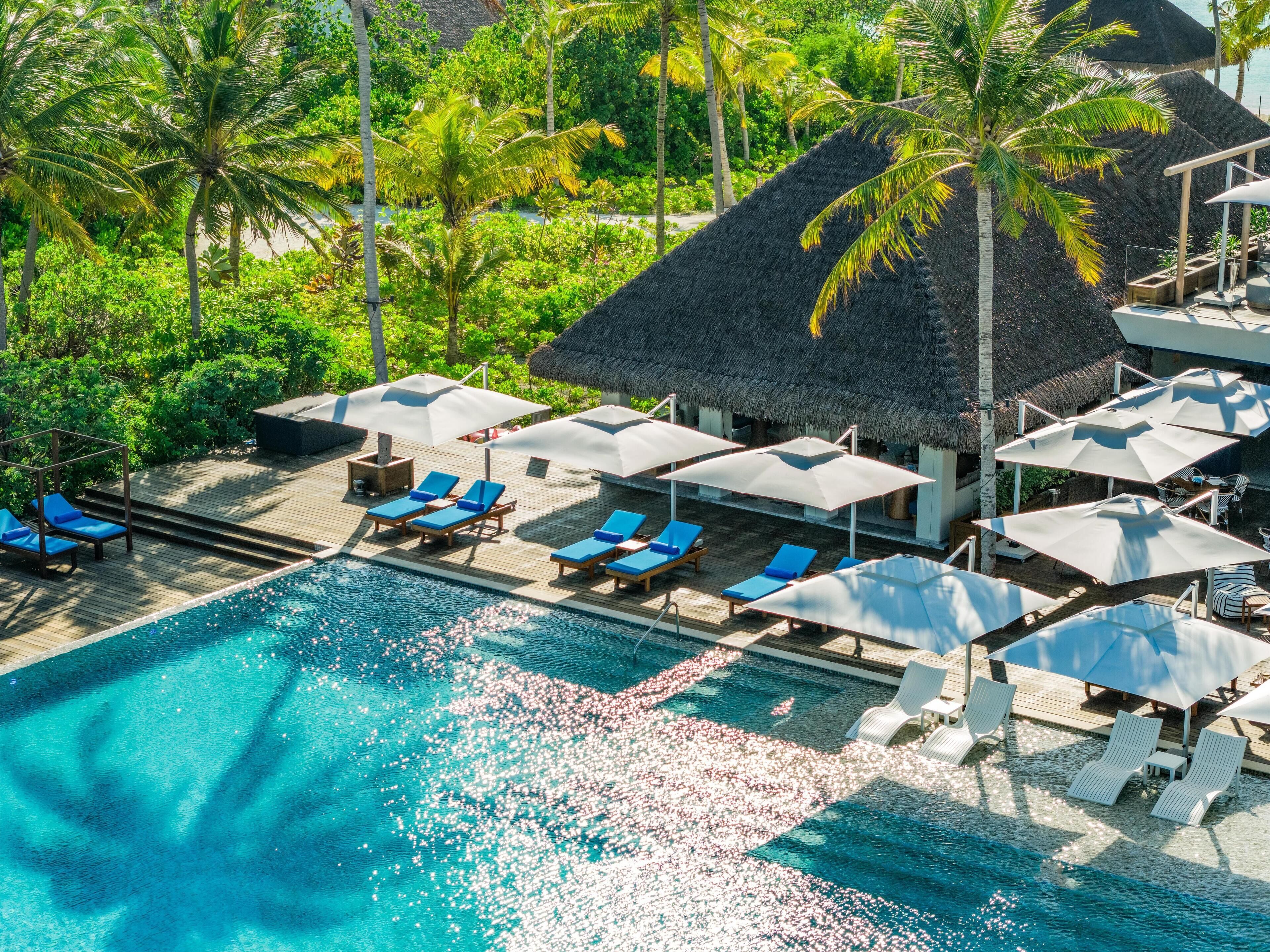 Vue aérienne d’un complexe hôtelier doté d’une piscine entourée de chaises longues et de parasols, de palmiers et de bâtiments à toits de chaume.