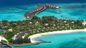 Luchtbeeld van een resort met bungalows boven water, een strand en helder turquoise water.